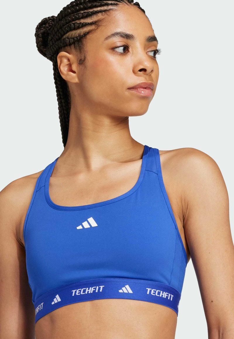 Blue sports bra with a smooth texture, wide straps, and a white logo. The band features "TECHFIT" in contrasting blue. Rounded neckline.