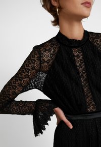 Robe en dentelle noire avec manches transparentes ornées de motifs floraux complexes, corsage plissé et ceinture en satin à la taille. Détail texturé dans le tissu.