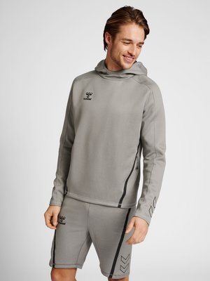 Hombre con sudadera con capucha gris de hummel y pantalones cortos a juego, de pie y mirando hacia abajo con una sonrisa sobre un fondo liso.