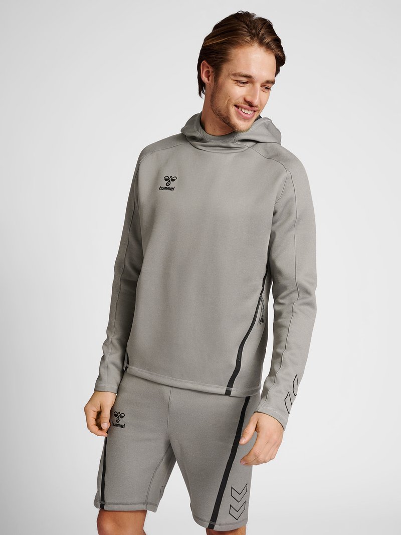 Homme portant un sweat à capuche gris hummel et un short assorti, debout et regardant vers le bas avec un sourire sur un fond uni.
