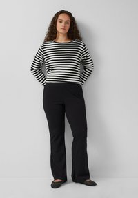 Gestreiftes schwarz-weißes Langarmoberteil mit gerippter Textur, kombiniert mit schwarzen Schlaghosen. Die Schuhe haben Nietenakzente.