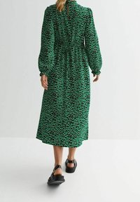 Vihreä kuvioitu pitkä mekko, jossa on pitkät pussihihat ja vyötäröä korostava istuvuus. Mekossa on takana kiinnitys ja laskeutuva kangas. Yhdistetty mustiin sandaaleihin.