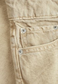 Tessuto denim beige con un sottile motivo a spina di pesce, dotato di bottoni a pressione in metallo argentato e dettagli cuciti su una tasca a toppa.