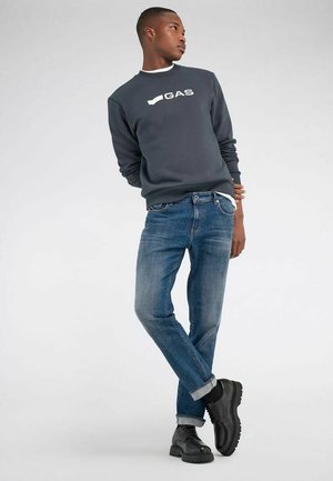 Junger Mann, der einen dunklen Pullover mit dem "GAS"-Logo, blaue Jeans mit aufgekrempelten Bündchen und schwarze klobige Schnürschuhe trägt, steht mit übereinandergeschlagenen Beinen.