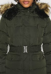 Femme portant une veste d'hiver matelassée vert foncé avec capuche bordée de fourrure, col montant, poches avant, et taille élastique ceinturée avec une boucle argentée.