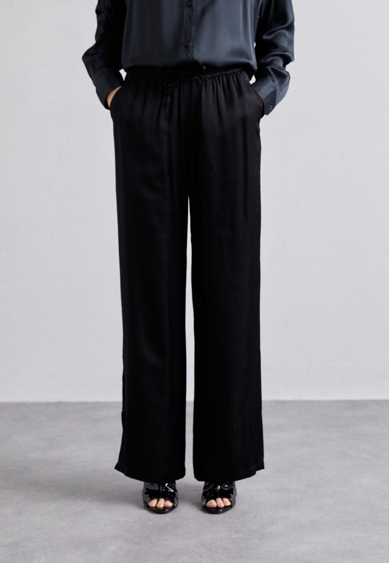 Svarta vida byxor med elastisk midja, med två sidofickor och ett mjukt, lätt tyg. Tillsammans med öppna sandaler.