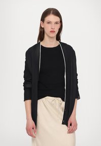 Black cardigan with a lightweight, sheer texture, open front, and long sleeves, layered over a fitted black top and light beige skirt.