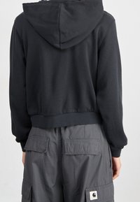 Person som bär en svart cropped hoodie och grå cargobyxor med stora ficklock, visad från baksidan mot en enfärgad bakgrund.