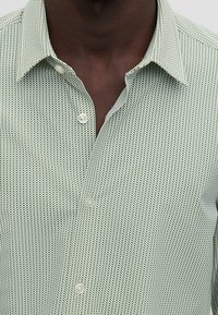 Groen patroonhemd met een smalle kraag, knopen en een gestructureerde stof met kleine geometrische vormen in donkerder groen.