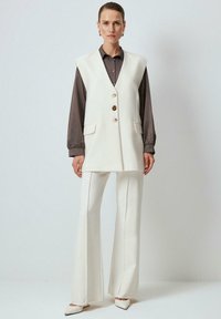 Chaleco de lana blanco con tres botones, dos bolsillos, superpuesto a una blusa marrón, combinado con unos pantalones acampanados blancos y zapatos de punta blanca.