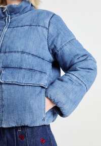 Young person wearing a blue padded denim jacket with hands in side pockets and dark denim pants with red butterfly embroidery.