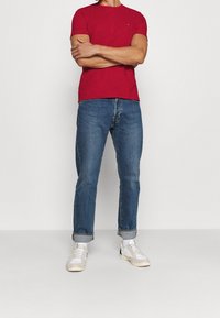 T-shirt rossa a maniche corte con scollatura a girocollo, abbinata a jeans blu risvoltati e sneakers bianche. Design semplice, vestibilità casual e linee pulite.