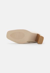Zapato de tacón beige con una parte superior suave y brillante, un tacón robusto y una suela de goma texturizada que presenta ranuras horizontales para un mejor agarre.