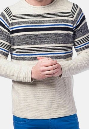 Adulte portant un pull en tricot crème avec des rayures horizontales noires, grises et bleues, se tenant les mains jointes devant, porte un pantalon bleu.
