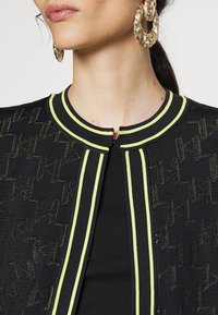 Czarna kurtka z teksturowanym wzorem, neonowożółtym obszyciem i okrągłym dekoltem. W zestawie duże, złote kolczyki-koła.