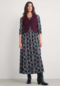 Burgundski prsluk preko uzorčaste haljine dugih rukava u tirkiznoj boji s tamnoljubičastim cvjetnim uzorkom. Manekenka nosi crne čizme s potpeticama.