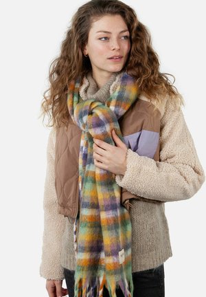Multicolored plaid scarf featuring green, yellow, purple, and white patterns, made of soft fabric with fringed edges, worn over a beige sweater.
