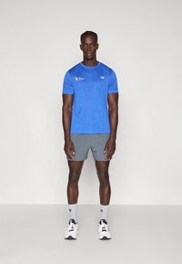 Zils sporta krekls ar īsām piedurknēm, pelēki šorti, balti sporta apavi un pelēkas zeķes. Modelis stāv pret vienkāršu fonu.