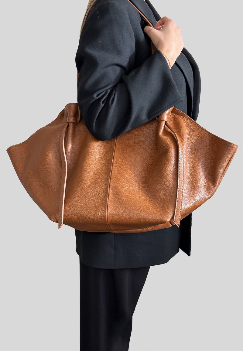 Personne en tenue noire tenant un grand sac à main en cuir marron sur l'épaule sur fond uni.