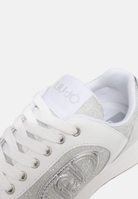 Sneaker bianca con dettagli in glitter argento, rifiniture a texture, punta rotonda, chiusura con lacci e logo "LIU•JO" sulla linguetta.