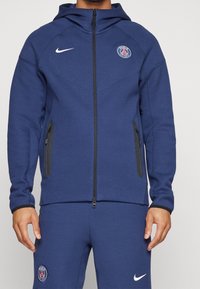 Veste à capuche bleu marine avec une fermeture éclair frontale, logo Nike et emblème de Paris ; dotée de poches zippées noires et d'un tissu texturé.
