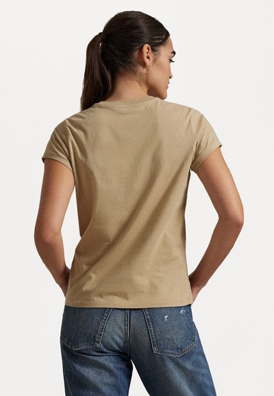 Beżowa bawełniana koszulka z krótkim rękawem i luźnym krojem. Tylna strona ma prosty wzór z minimalną ilością szwów. Założona z przetartymi dżinsami.