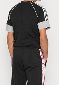 Maglietta sportiva nera con maniche grigie e strisce bianche lungo le spalle. I pantaloni neri presentano strisce verticali rosse e bianche sui lati.