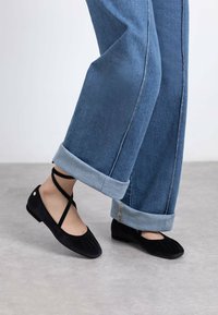 Bailarinas de ante negro con un diseño plisado, que cuentan con tiras cruzadas y una suela plana, combinadas con unos vaqueros de mezclilla de corte recto y puños en azul claro.