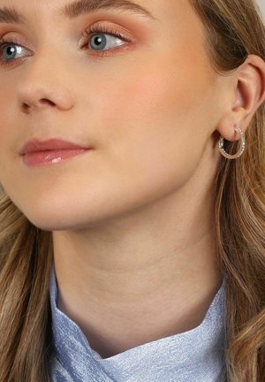 Söljda hoops örhängen med en texturerad yta och små detaljer. Modellen bär en ljusblå, silkig topp. Hudtonen verkar neutral.