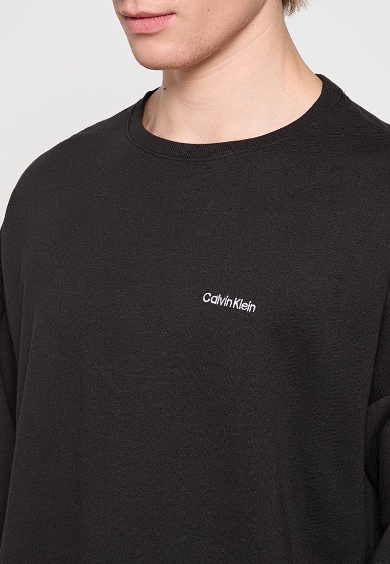 T-shirt a maniche lunghe in cotone nero con collo rotondo e logo bianco "Calvin Klein" sul lato sinistro del petto. Texture liscia.