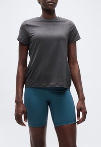 Άτομο που φοράει εφαρμοστό σκούρο γκρι κοντομάνικο μπλουζάκι και αθλητική βερμούδα ψηλόμεση σε χρώμα τυρκουάζ, στέκεται μπροστά σε ένα απλό φόντο.