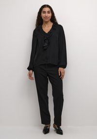 Black long-sleeve blouse with ruffled neckline, paired with tailored black trousers and pointed black shoes, against a plain background.