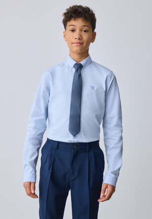 Niño con cabello corto y rizado lleva una camisa de vestir azul claro, corbata azul oscuro y pantalones de vestir azul oscuro, de pie frente a un fondo sencillo.