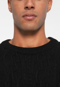 Schwarzer Strickpullover mit einem strukturierten Muster und einem gerippten runden Ausschnitt, der eine Nahaufnahme des Kragens und der oberen Brust zeigt.