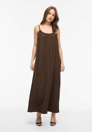Jeune femme aux longs cheveux bruns portant une robe maxi marron texturée sans manches et des talons noirs ouverts à l'avant, debout devant un fond blanc.