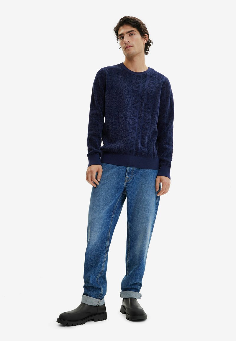 Pull en tricot bleu marine avec un motif texturé, col rond et poignets côtelés, associé à un jean bleu clair à coupe décontractée et des bottines noires.