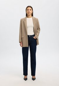 Beige blazer with structured shoulders over a white shirt, paired with dark blue jeans and black pointed-toe heels. Smooth fabric and tailored fit.
