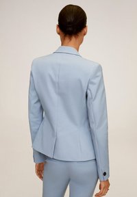 Blazer bleu clair sur mesure avec une silhouette ajustée ; comporte des épaules structurées, une fente unique et des boutons noirs aux poignets.