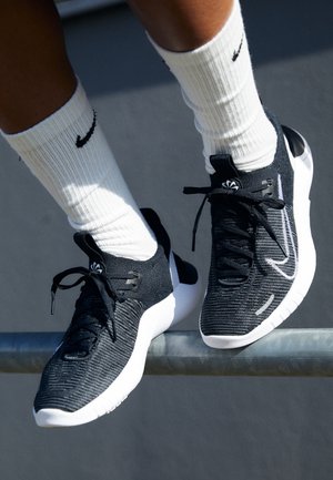 Jambes portant des chaussettes blanches Nike et des baskets Nike noires et blanches reposant sur une barre métallique sur un fond sombre.