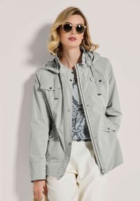 Hellegraue wasserdichte Jacke mit Kapuze, Frontreißverschluss, Druckknöpfen und zwei Brusttaschen; getragen über einem gemusterten Oberteil und cremefarbenen Hosen.