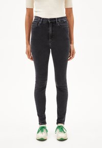 Jeans skinny en denim noir avec une taille haute, comportant des poches avant et une texture lisse. Portés avec des baskets blanches et des lacets verts.