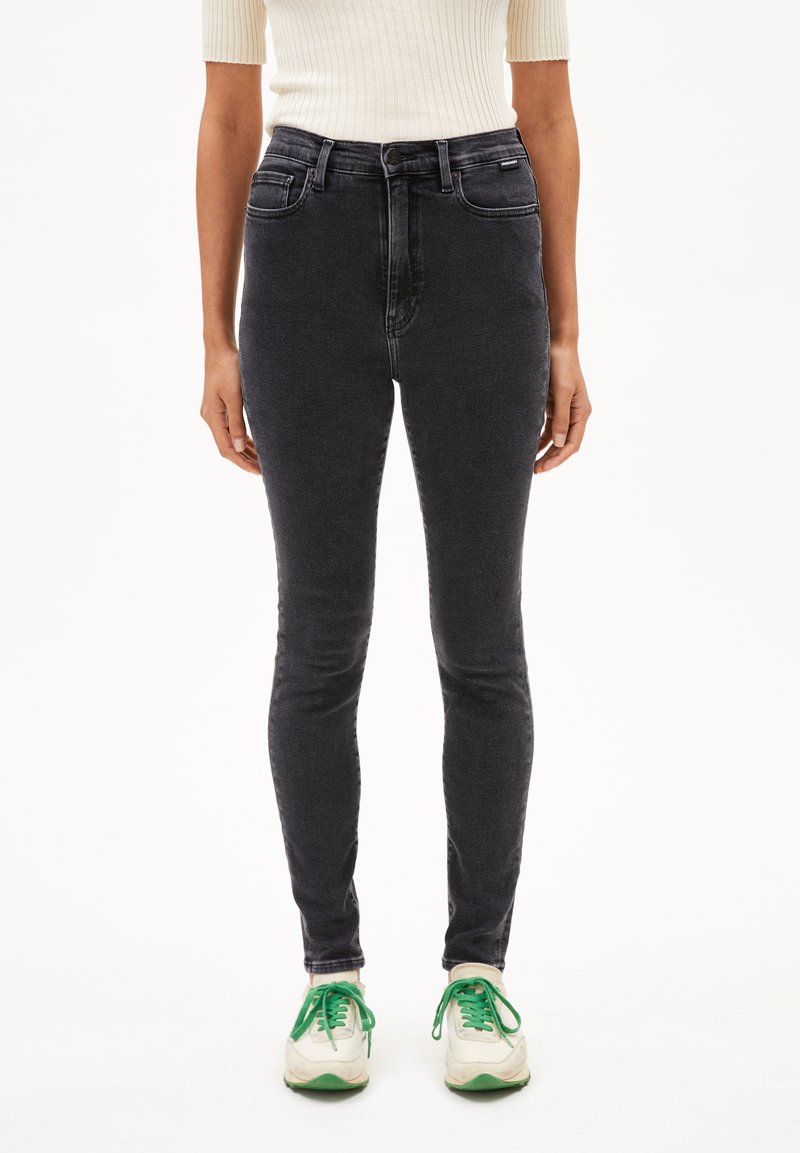 Jeans skinny en denim noir avec une taille haute, comportant des poches avant et une texture lisse. Portés avec des baskets blanches et des lacets verts.