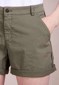 Pantaloni corti verde oliva realizzati in cotone morbido con una texture leggermente spiegazzata. Presentano un orlo arrotolato e tasche laterali, fissate con un bottone.