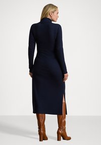 Abito lungo navy a maniche lunghe con colletto a imbuto, fessure laterali, silhouette aderente e texture morbida. Abbinato a stivali marroni alti fino al ginocchio con tacco.