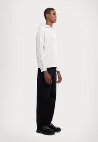 Hanorac alb cu un model, dotat cu un glugă cu șnur, purtat cu pantaloni largi negri și pantofi negri. Textură fină și croială lejeră.