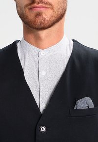 Schwarze Weste über einem weißen Hemd mit Kragen und gepunktetem Muster. In der Vorderasche der Weste ist ein graues Taschentuch sichtbar. Glatte Stoffoberfläche.