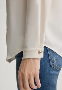 Blusa beige chiaro con maniche lunghe, dotata di chiusura a bottone dorato ai polsini. Texture liscia e vestibilità leggermente ampia.