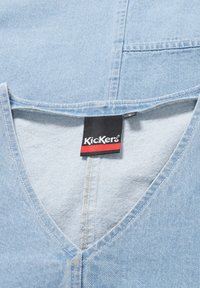 Top in denim blu chiaro con scollatura a V e un'etichetta che riporta la scritta "Kickers" in nero e rosso. Presenta cuciture visibili e un'etichetta di taglia.
