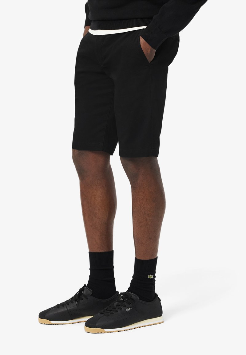 Shorts noirs en coton lisse, avec deux poches latérales et une coupe légèrement ajustée. Associés à des baskets noires et des chaussettes mi-mollet.