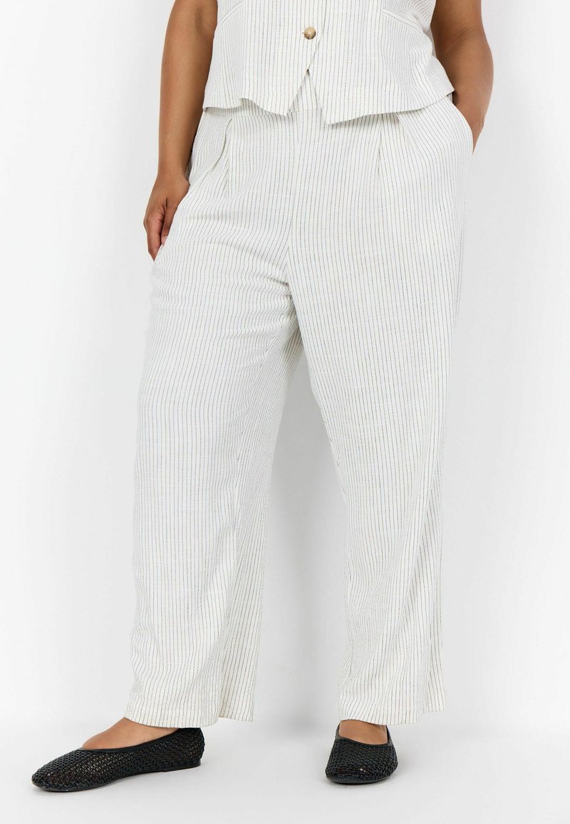 Nő fehér csíkos, bő szárú nadrágot és fekete fonott lapos cipőt visel, zsebében a kezével, sima háttér előtt áll.